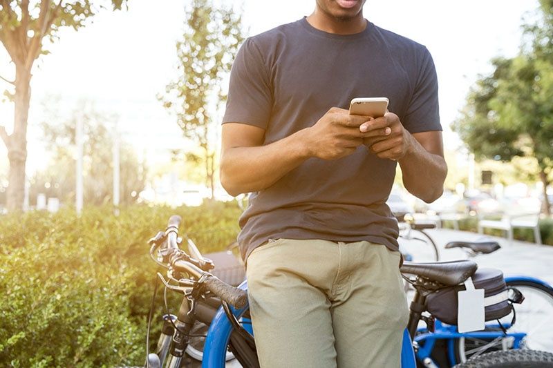 man leaning on a bicycle on a sunny day and interacting with his cell phone